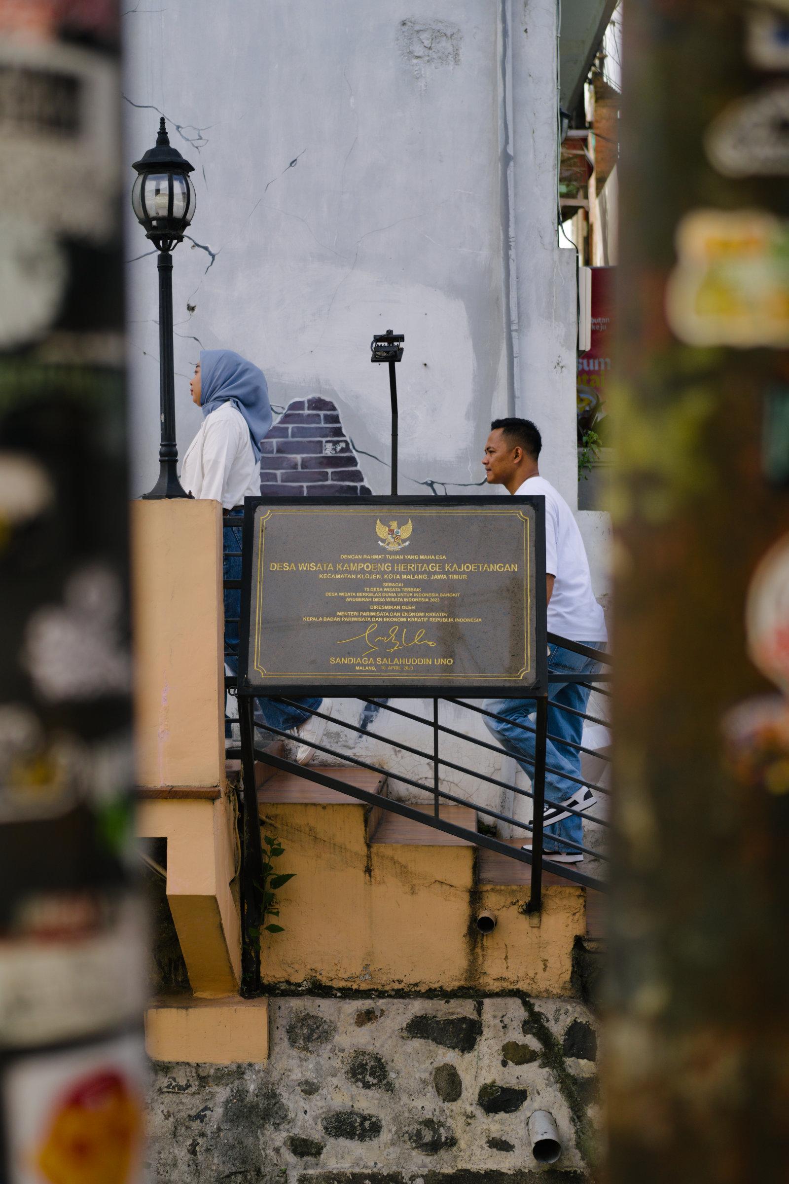 The inauguration plaque of the renovated Kajoetangan area, seen through the space between two utility poles. A couple can be seen walking behind it.