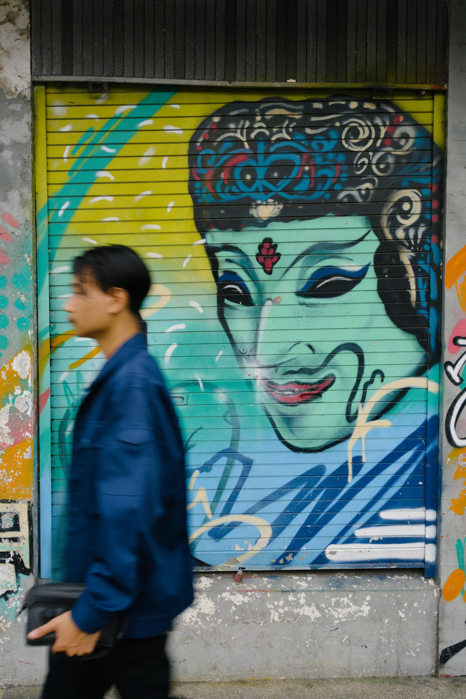 Someone walking past a wall with an drawing of stylized wayang face on it. The framing makes it look like the drawing is watching him.